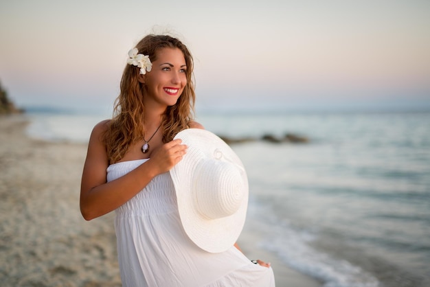 Ritratto di una giovane e bella donna sorridente in abito bianco che cammina sulla spiaggia e tiene in mano un cappello estivo bianco.