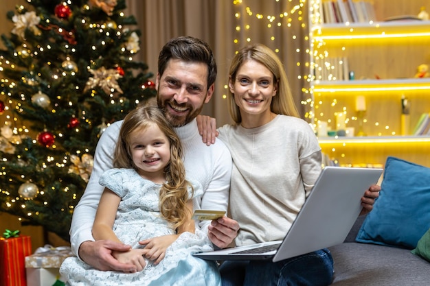 Ritratto di una giovane famiglia seduta sul divano di casa vicino all'albero di natale con laptop e