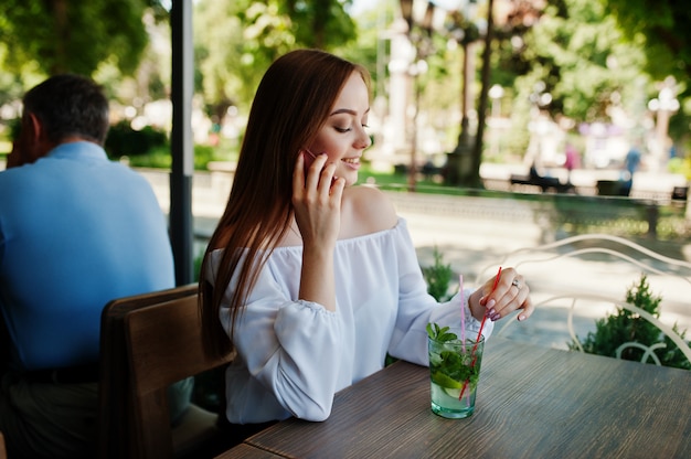 Ritratto di una giovane imprenditrice attraente in possesso di un cocktail mojito e parlando al telefono in un accogliente bar accanto al parco.