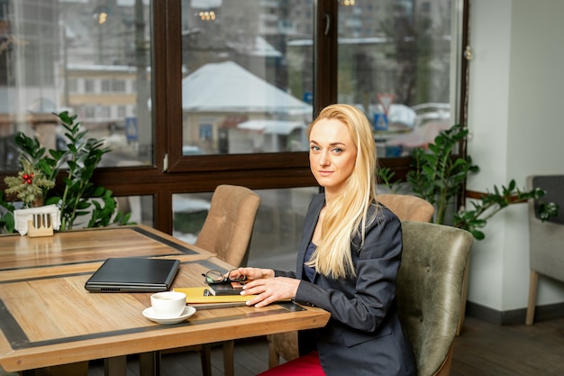 Ritratto di una giovane imprenditrice caucasica con documenti e laptop al tavolo guardando la telecamera nella caffetteria