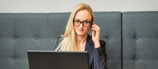 Ritratto di una giovane imprenditrice caucasica in bicchieri chiama su uno smartphone con un laptop al tavolo