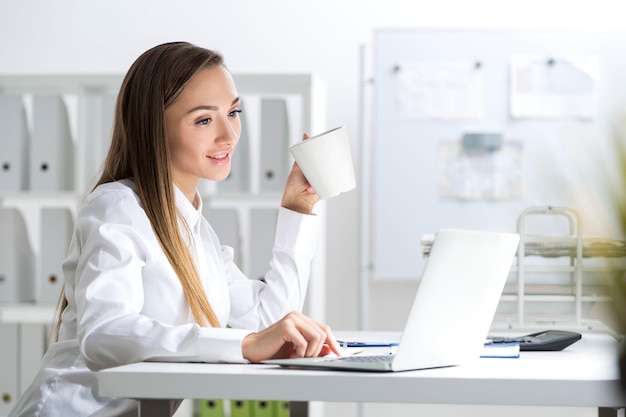 Ritratto di una giovane imprenditrice che indossa una camicia bianca e seduta in un ufficio. Tiene in mano la sua tazza, guarda lo schermo di un laptop e preme un tasto