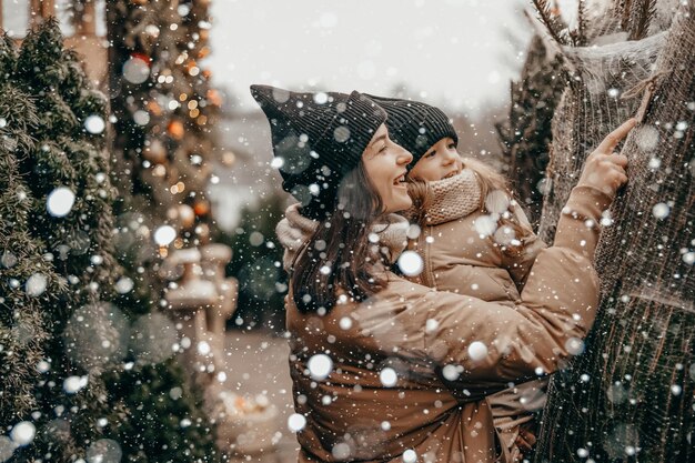 Ritratto di una giovane madre con la figlia in braccio sorridono e si rallegrano per la nevicata Scegliere e acquistare un albero di Natale al mercatino di Natale