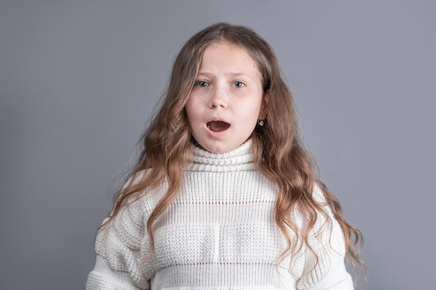 Ritratto di una giovane ragazza attraente con lunghi capelli biondi fluenti in un maglione bianco sorpreso stupire su uno sfondo grigio studio. Posto per il testo. Copia spazio.