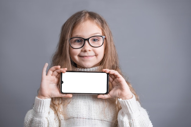 Ritratto di una giovane ragazza attraente con lunghi capelli biondi fluenti in una stretta maglione bianco mostra un telefono cellulare con uno schermo vuoto bianco su uno sfondo grigio studio. Posto per il testo. Copia spazio.