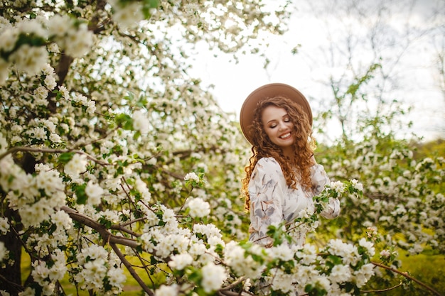 Ritratto di una giovane ragazza con i capelli ricci in piedi nel mezzo di un albero in fiore circondato da tanti fiori bianchi. Donna che indossa un cappello beige e abito bianco in una luminosa giornata di primavera.