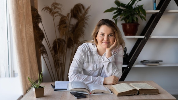 Ritratto di una giovane ragazza consulente seduta a un tavolo di legno con libri e libri di testo in ufficio vicino alla finestra Una donna si prepara per una videoconferenza e legge letteratura