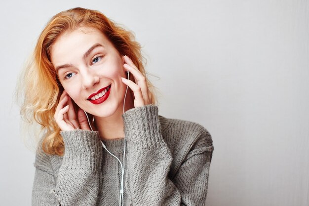 Ritratto di una giovane ragazza dai capelli rossi su uno sfondo grigio con un maglione che ascolta musica con le cuffie