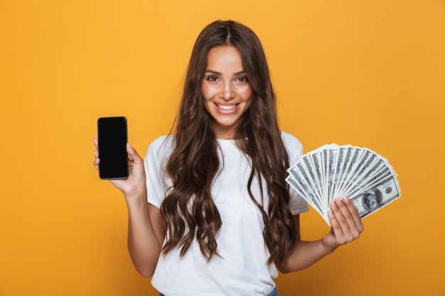 Ritratto di una giovane ragazza felice con lunghi capelli castani in piedi sopra il muro giallo, tenendo in mano banconote, mostrando il telefono cellulare con schermo vuoto