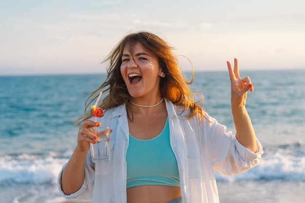 Ritratto di una giovane ragazza felice con un cocktail in mano su uno sfondo di mare bellissimo Ragazza che si diverte durante le vacanze estive La ragazza mostra la pace con due dita Libertà e felicità