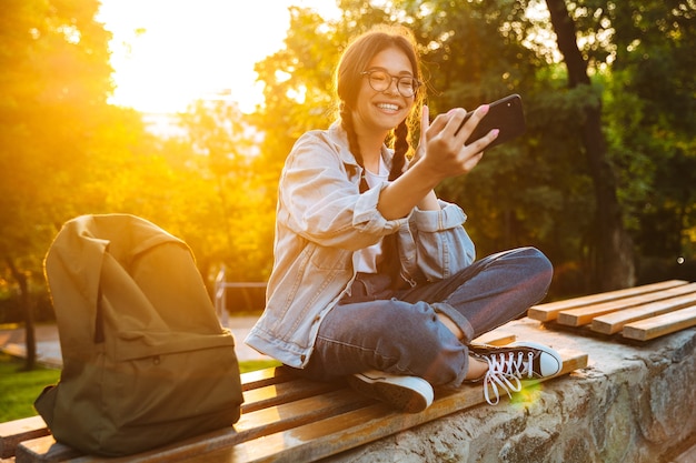 Ritratto di una giovane studentessa allegra e carina che indossa occhiali da vista seduta su una panchina all'aperto nel parco naturale con una bella luce solare utilizzando il telefono cellulare parlando prendere un selfie