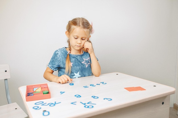 Ritratto di una graziosa bambina seduta a un tavolo bianco e che guarda pensieroso i numeri mentre pensa alla risposta di un'equazione di esempio. Concetto di matematica. foto con rumore