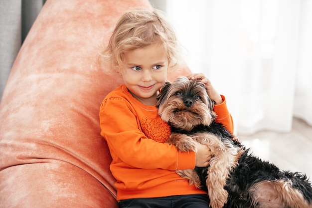 Ritratto di una graziosa bambina seduta su una sedia arancione con un cane in braccio che guarda di lato e sorride