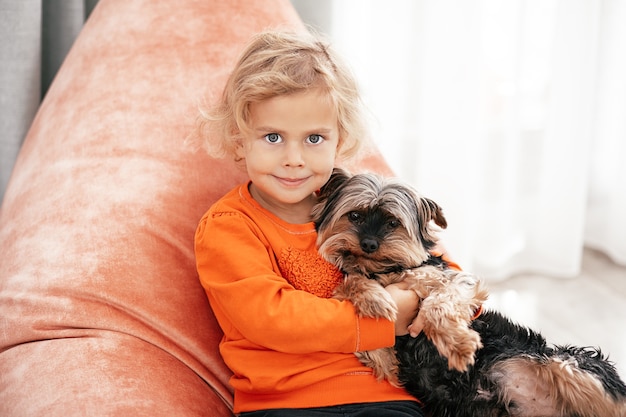 Ritratto di una graziosa bambina seduta su una sedia arancione con un cane in braccio che guarda l'obbiettivo e sorride