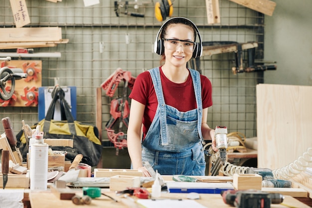 Ritratto di una graziosa carpentiere femminile con cuffie e occhiali che lavora con la cucitrice pneumatica durante la fabbricazione di mobili