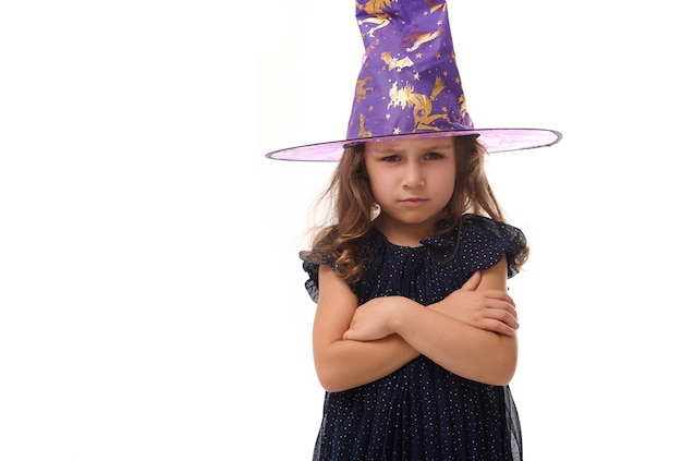 Ritratto di una graziosa strega arrabbiata ragazza arrabbiata che indossa un cappello da mago e vestita con un elegante abito di carnevale, che guarda l'obbiettivo in posa con le braccia incrociate su sfondo bianco, copia spazio. Halloween