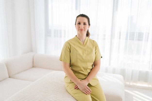 Ritratto di una massaggiatrice sorridente in uniforme seduta a posare sul divano guardando la telecamera con un'espressione amichevole sullo sfondo della finestra