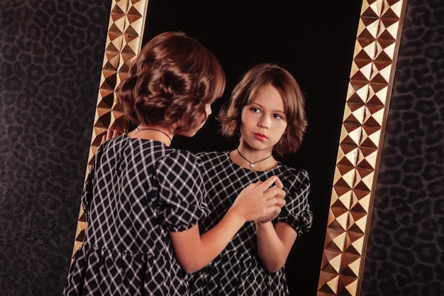 Ritratto di una ragazza adolescente abbastanza carina in un vestito elegante allo specchio in interni eleganti scuri del soggiorno. Emozioni dei bambini e in posa. Concetto di stile, moda e bellezza. Copia spazio per il sito