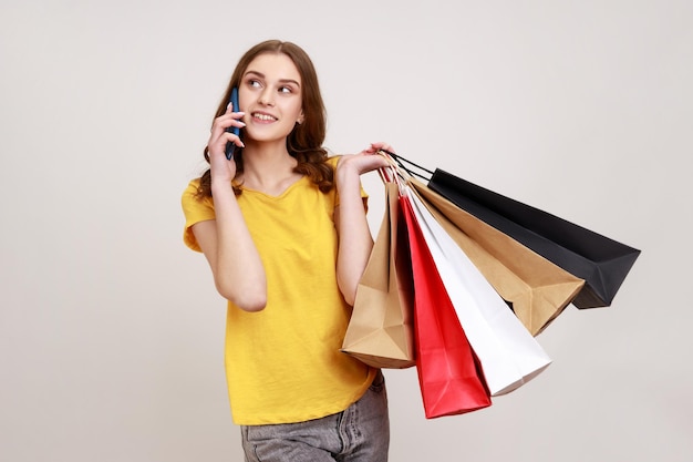 Ritratto di una ragazza adolescente abbastanza felice in maglietta gialla che tiene le borse della spesa, parla con entusiasmo al telefono cellulare, vantando l'acquisto nel negozio di moda. Studio indoor girato isolato su sfondo grigio