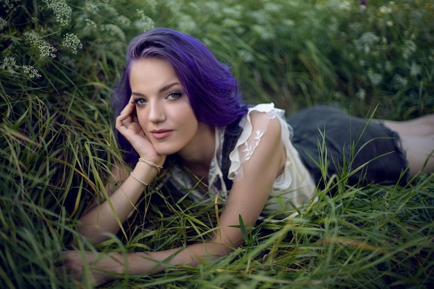 Ritratto di una ragazza adolescente con i capelli viola e un orecchino al naso sdraiato sull'erba in natura
