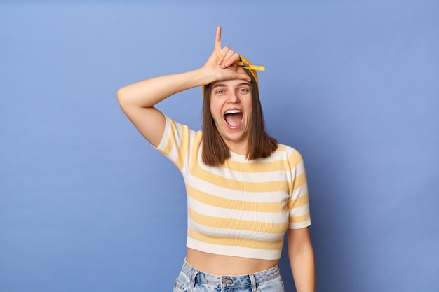 Ritratto di una ragazza adolescente eccitata estremamente felice che indossa una maglietta a righe e un berretto da baseball in piedi isolato su sfondo blu in piedi guardando la fotocamera che mostra il gesto con le dita