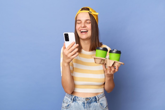 Ritratto di una ragazza adolescente felice e gioiosa che indossa una maglietta a righe e un berretto da baseball in piedi isolato su sfondo blu con in mano due tazze di caffè usa e getta e utilizza uno smartphone