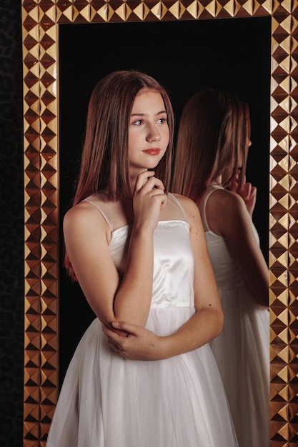 Ritratto di una ragazza adolescente piuttosto carina in un vestito elegante allo specchio nell'interno scuro ed elegante del soggiorno Emozioni dei bambini e posa Concetto di stile, moda e bellezza Copia spazio per il sito