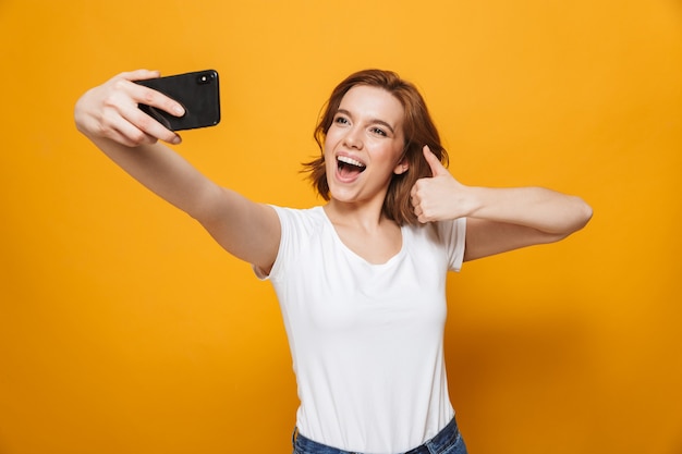 Ritratto di una ragazza adorabile felice in piedi isolata sul muro giallo, facendo un selfie, pollice in alto