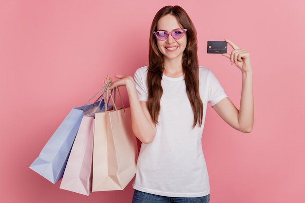 Ritratto di una ragazza affascinante per lo shopping che tiene la carta di credito delle borse del negozio su sfondo rosa