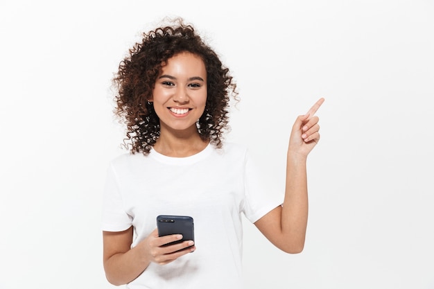 Ritratto di una ragazza africana casual piuttosto allegra in piedi isolata sul muro bianco, usando il telefono cellulare, puntando il dito lontano