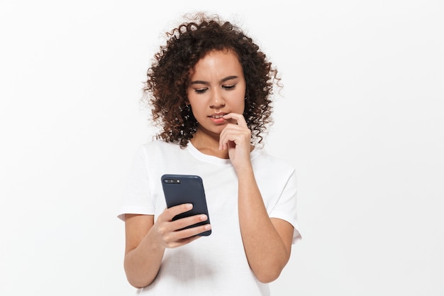 Ritratto di una ragazza africana casual piuttosto pensierosa in piedi isolata sul muro bianco, usando il telefono cellulare