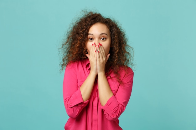 Ritratto di una ragazza africana scioccata in abiti casual rosa che copre la bocca con le mani isolate sul fondo della parete turchese blu in studio. Persone sincere emozioni, concetto di stile di vita. Mock up copia spazio.