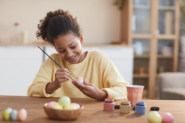 Ritratto di una ragazza afro-americana sorridente che dipinge a mano le uova di Pasqua mentre si gode la decorazione fai-da-te a casa, copia spazio