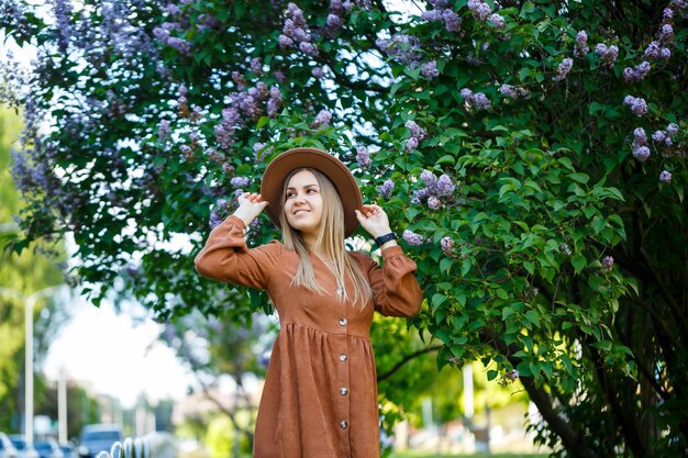 Ritratto di una ragazza alla moda in un cappello marrone e vestito su uno sfondo di albero di lillà in una giornata calda e soleggiata. Una giovane donna dall'aspetto europeo con un sorriso sul viso