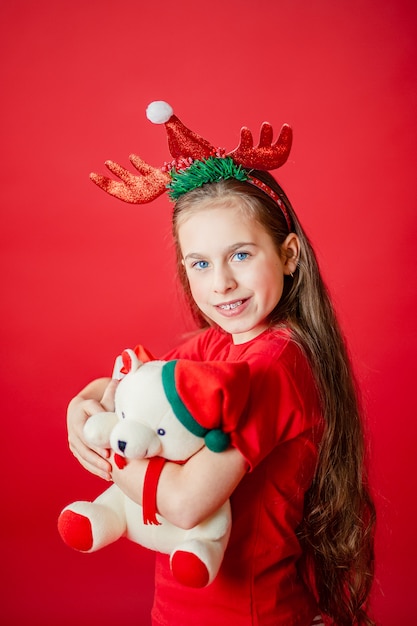 Ritratto di una ragazza allegra divertente con una benda di corna sulla sua testa che abbraccia un orsacchiotto in pigiama di Natale isolato
