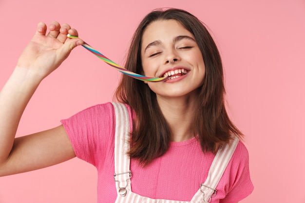 Ritratto di una ragazza allegra e affascinante che sorride e mangia marmellata multicolore isolata su un muro rosa