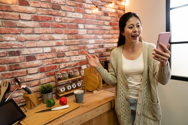 Ritratto di una ragazza amichevole positiva che ha una videochiamata che gesturing il segno ciao del palmo. giovane moglie di casa che mostra il suo nuovo condominio cucina muro di mattoni sul retro pronto a cucinare con verdure e tablet