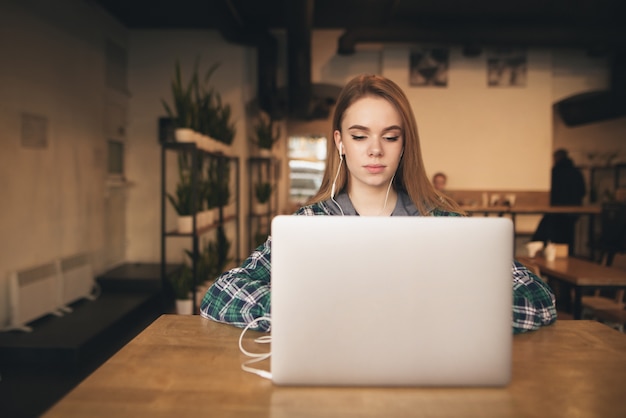 Ritratto di una ragazza attraente con un laptop nel caffè, ascolta la musica in cuffia e guarda lo schermo.