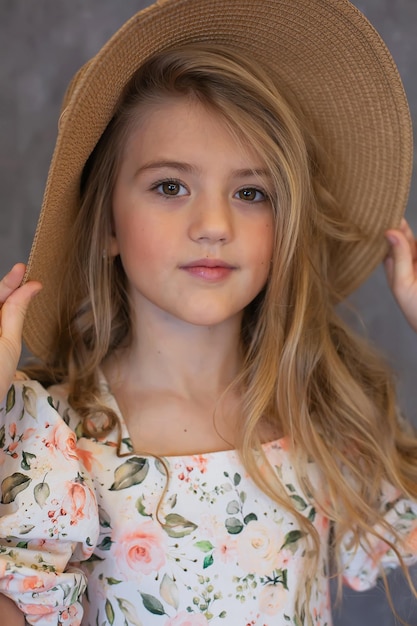 ritratto di una ragazza bionda felice dai capelli lunghi in un abito floreale e un cappello di paglia su uno sfondo grigio