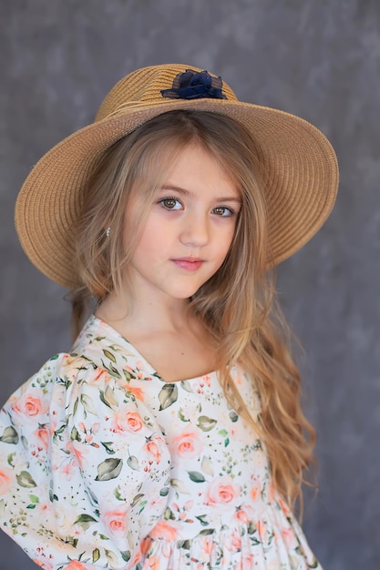 ritratto di una ragazza bionda felice dai capelli lunghi in un abito floreale e un cappello di paglia su uno sfondo grigio