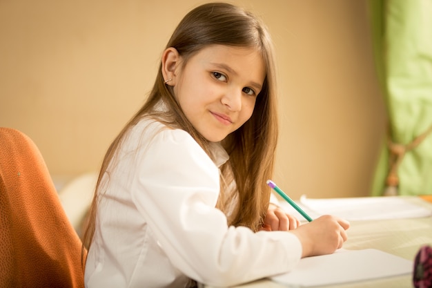 Ritratto di una ragazza bruna sorridente in camicia bianca seduta dietro la scrivania e che fa i compiti