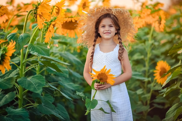 Ritratto di una ragazza carina che ride in abiti leggeri e un grande cappello di paglia al tramonto su un campo di girasoli. tiene in mano un grande fiore