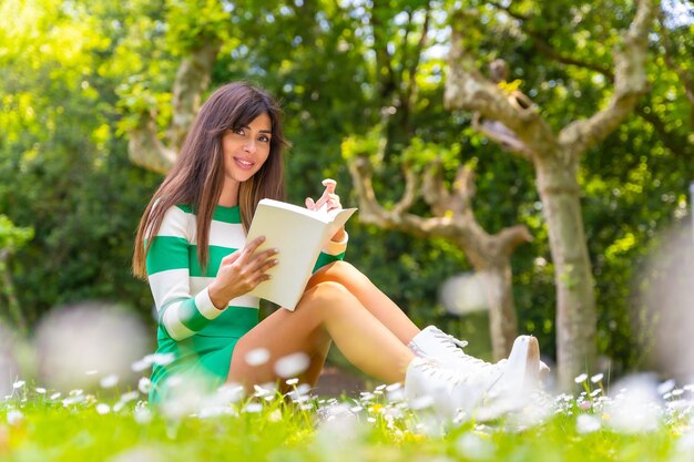 Ritratto di una ragazza caucasica bruna che legge un libro in natura seduta sull'erba che indossa un maglione verde e bianco guardando la telecamera