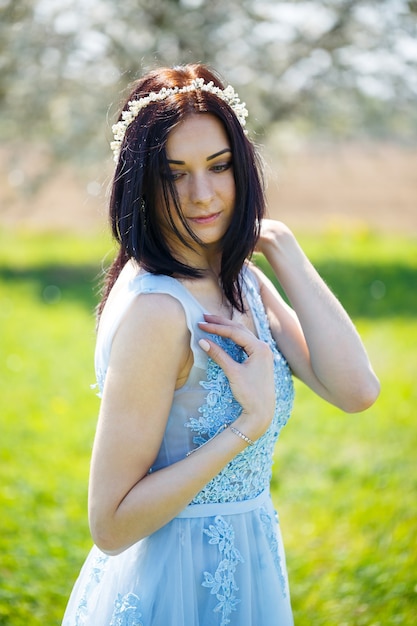 Ritratto di una ragazza con un abito lungo azzurro estivo adornato tra i capelli contro un albero in fiore.