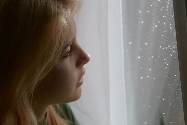 ritratto di una ragazza di 18 anni con i capelli biondi vicino alla finestra in un giorno di pioggia
