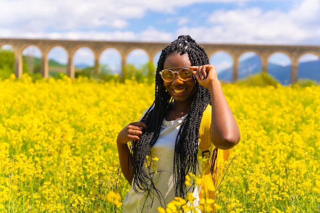 Ritratto di una ragazza di etnia nera con trecce e occhiali da sole viaggiatore in un campo di fiori gialli