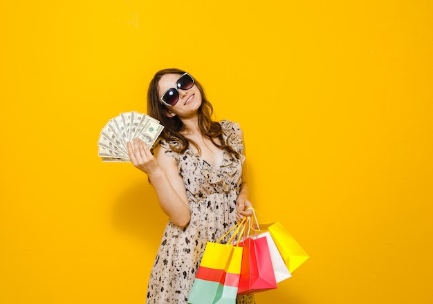 Ritratto di una ragazza felice con gli occhiali da sole che mostrano il mazzo di banconote dei soldi mentre gesto e che stanno e che tengono i sacchetti della spesa