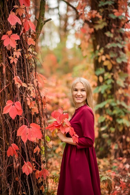 Ritratto di una ragazza in un abito bordeaux in una foresta autunnale. Foglie rosse e arancioni tutt'intorno..