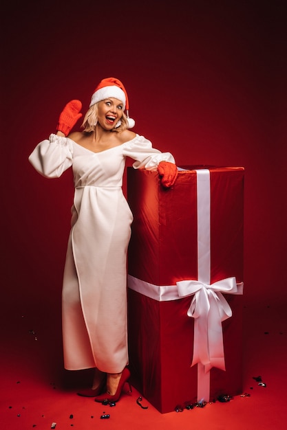 Ritratto di una ragazza sorridente in abito bianco e cappello da Babbo Natale con un enorme regalo di Natale su sfondo rosso.