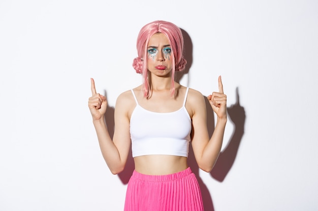 Ritratto di una sciocca ragazza delusa con una parrucca rosa, che punta le dita in alto e tiene il broncio, lamentandosi di qualcosa di brutto, in piedi su sfondo bianco.